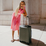 Smiling woman in a pink dress posing with a durable, large suitcase from Samsonite in a sunny outdoor setting.