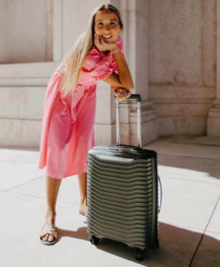 Smiling woman in a pink dress posing with a durable, large suitcase from Samsonite in a sunny outdoor setting.